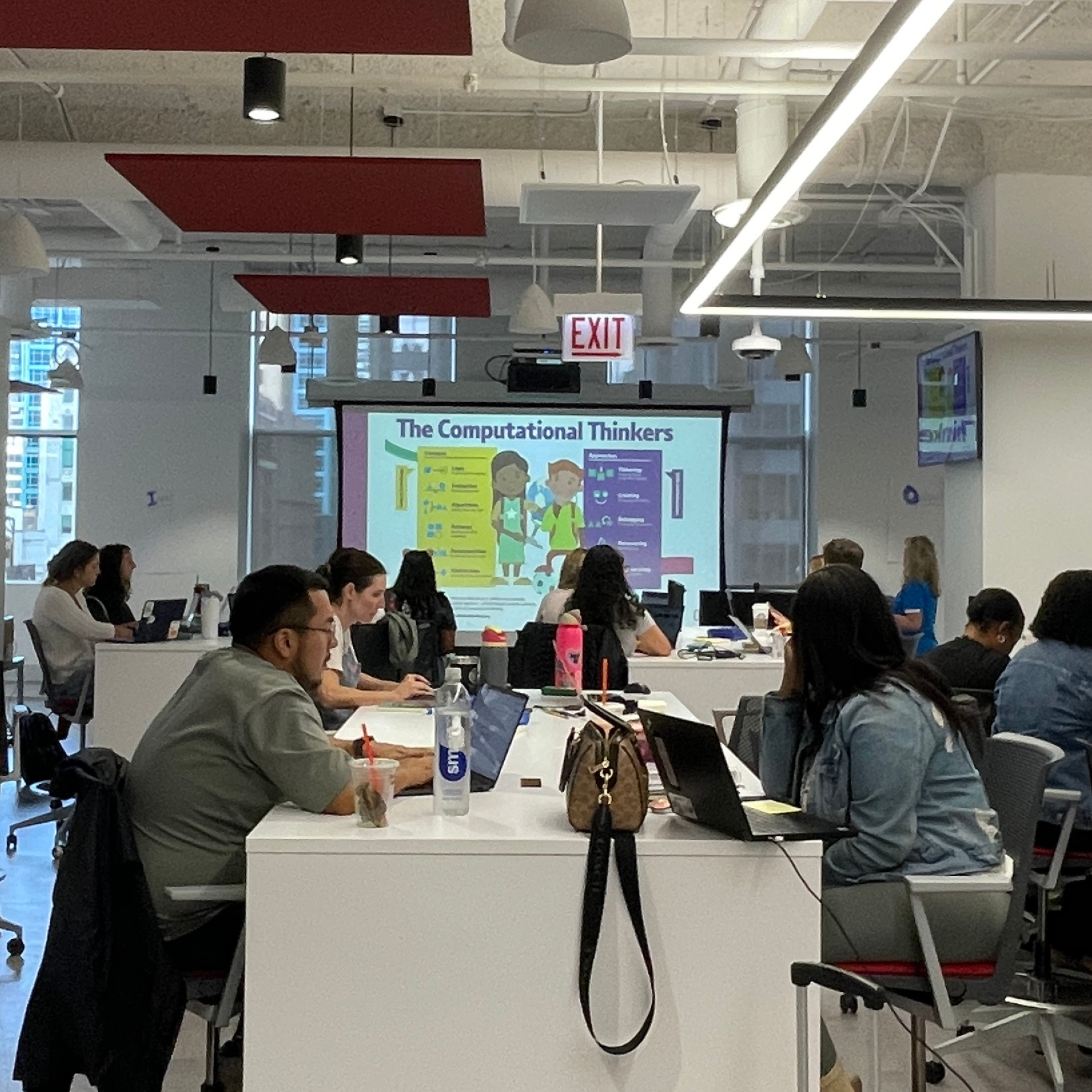 A group of educators gathered around a table as they observe a projected slide that reads "The Computational Thinker"