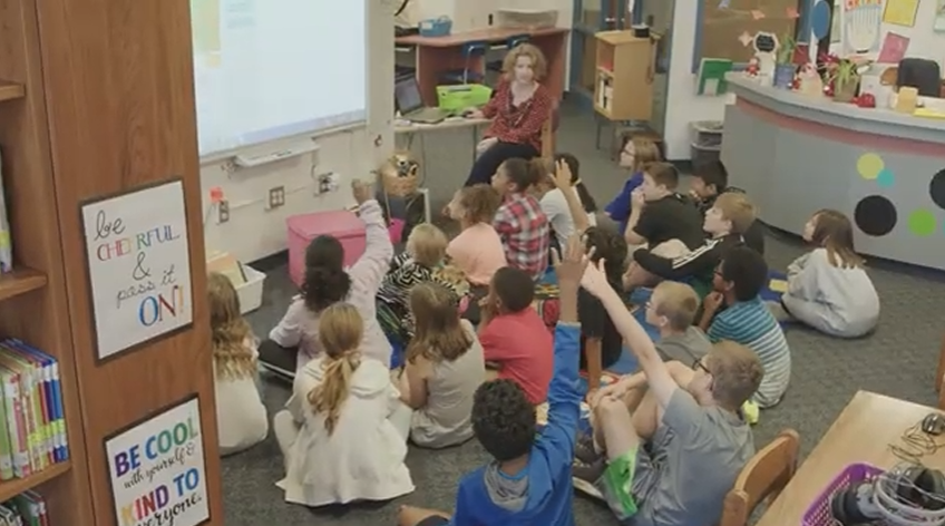 An educator sitting in front of a large group of children sitting on the floor. A few students have their hands raised to answer a question.