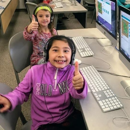 Two children sitting in front of computers looking at the person photographing them and smiling and giving a thumps up